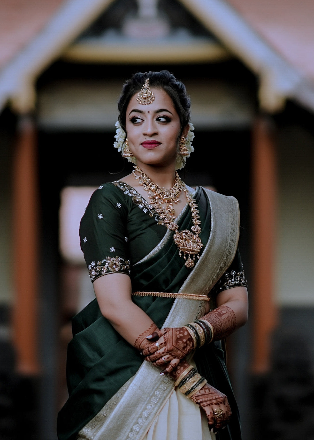 Woman in traditional attire with jewelry and makeup, standing in front of a blurred architectural background.