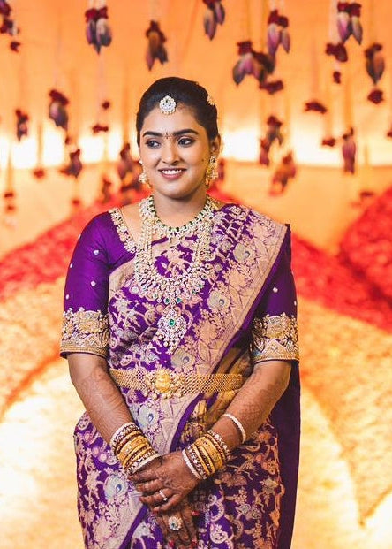 Bride in a traditional purple saree with gold jewelry against a decorative background