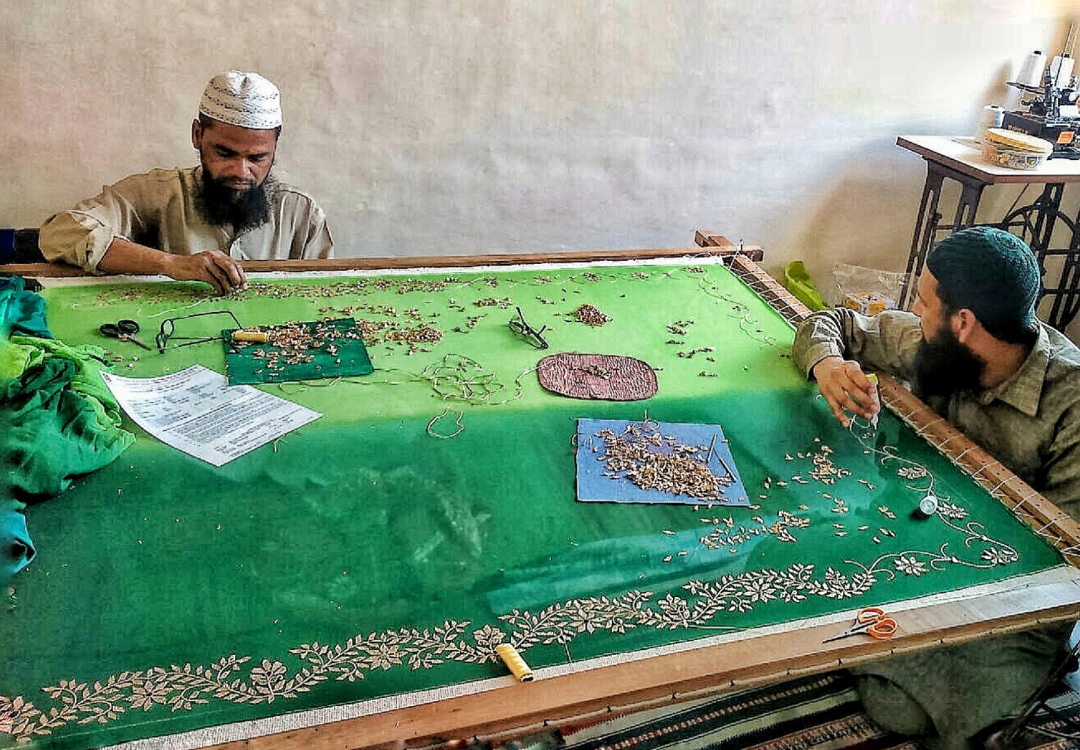 Two men working on an embroidery project with a large green fabric and various tools.