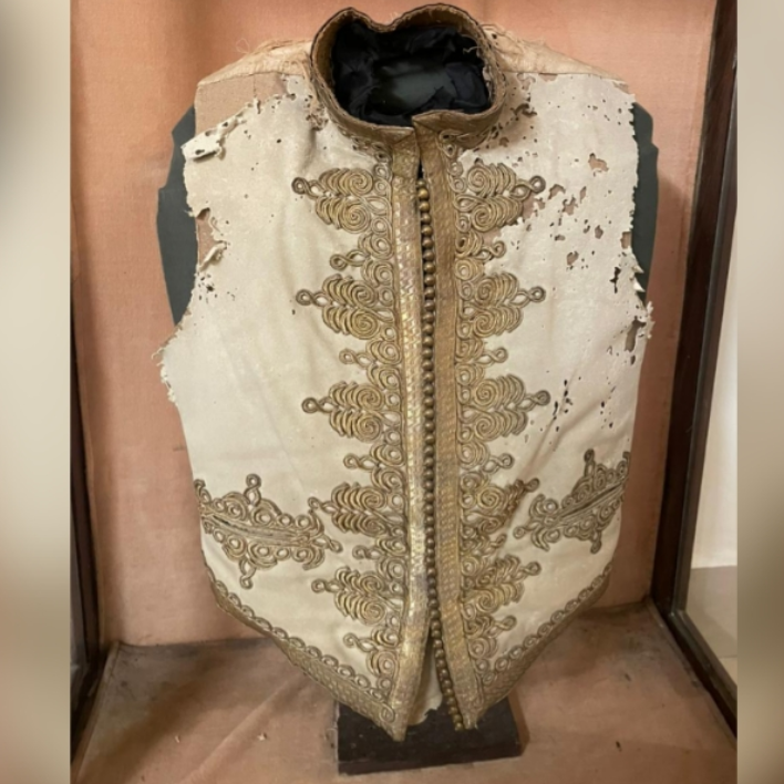 Decorative vest with intricate gold embroidery on a wooden stand.